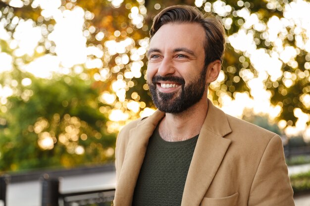 handsome caucasian bearded adult man in jacket smiling and walking in park