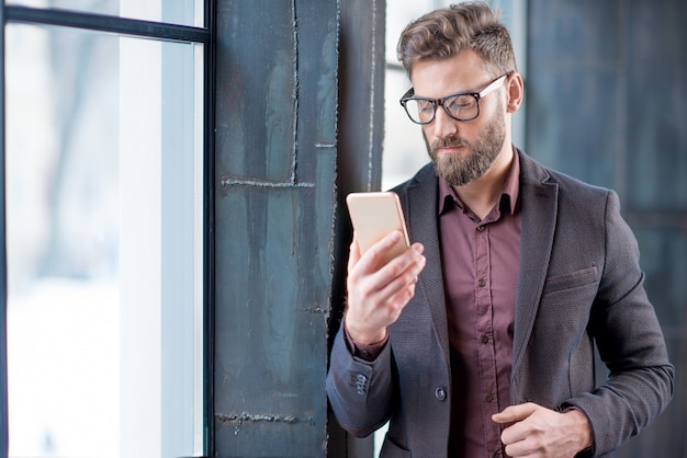 Handsome caucasian businessman dressed in the suit reading with his smart phone near the window in the loft interior studio