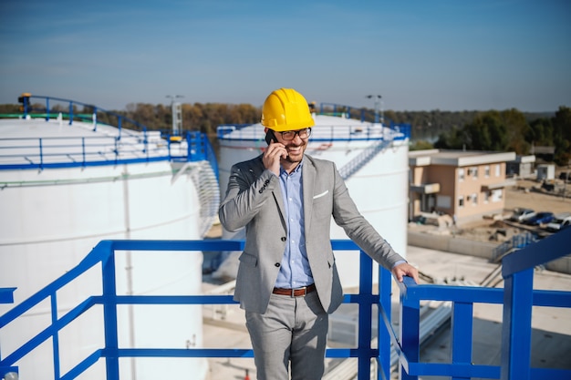 Uomo d'affari caucasico bello in vestito e con il casco sulla testa che parla sul telefono mentre stando all'aperto. sullo sfondo si trovano i serbatoi dell'olio.