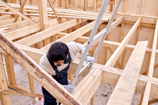 Handsome caucasian confident male, professional hardworking contractor in work uniform go down the ladder after finished work in roof, in wooden house building site alone