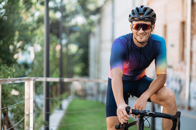 Handsome caucasian man in colorful sport clothes helmet and glasses smiling on camera while sitting on black bike Outdoors training of cyclist