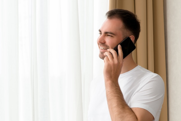 Handsome Caucasian man looking through window in hotel while speaking on smartphone