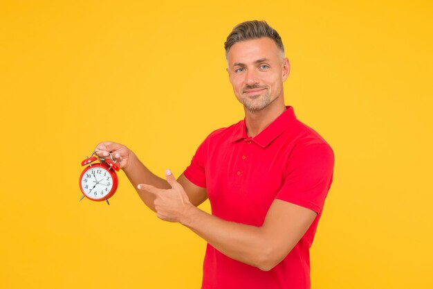 Handsome caucasian man of middle age point finger at mechanical alarm clock yellow background, time.