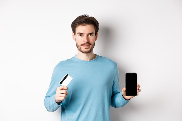 Handsome caucasian man showing empty smartphone screen and plastic credit card, standing over white background.