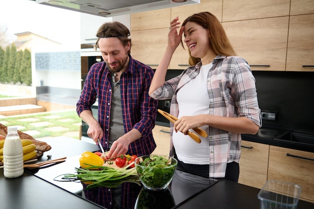 Handsome caucasian man slicing vegetables, preparing healthy\
vegan food standing next to his beloved pregnant woman in the home\
kitchen, enjoying happy time together. happiness, tenderness,\
maternity