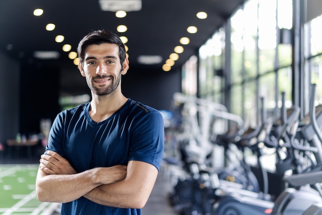 Handsome Caucasian man with beard in blue color sportswear standing and crossing arms in gym or fitness club.