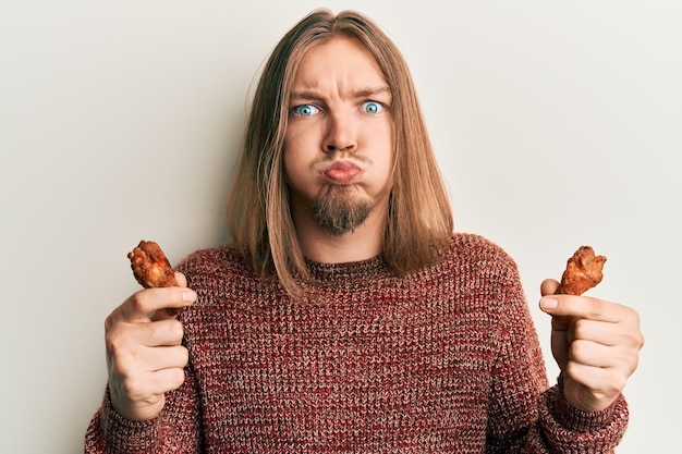 写真 変な顔で頬を膨らませて手羽先を食べる長い髪のハンサムな白人男性。口が空気で膨らみ、空気をキャッチします。