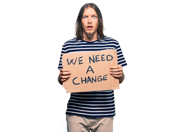 Handsome caucasian man with long hair holding we need a change banner scared and amazed with open mouth for surprise disbelief face