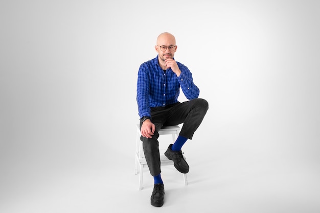 Handsome caucasian man without hair in blue shirt, black trousers and blue socks sits on a long chair and thinks about something