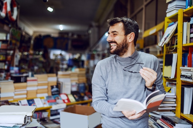 Красивый кавказский усмехаясь человек стоя в книжном магазине с книгой в руках и смотря прочь.