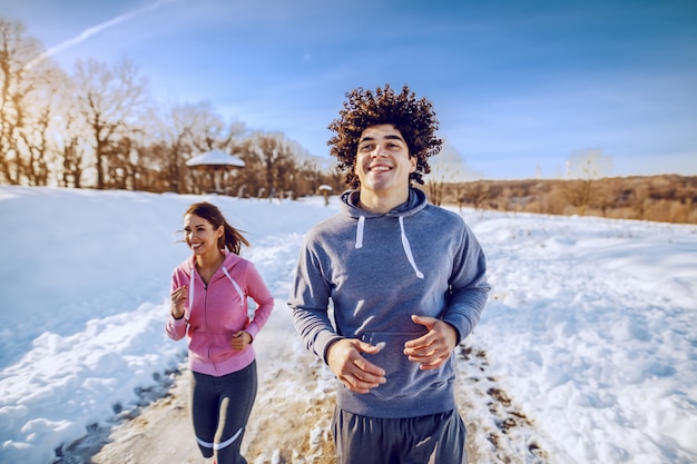 Uomo sorridente caucasico bello con capelli ricci e in abiti sportivi che corrono in natura con il suo amico femminile. orario invernale. concetto di fitness all'aperto.