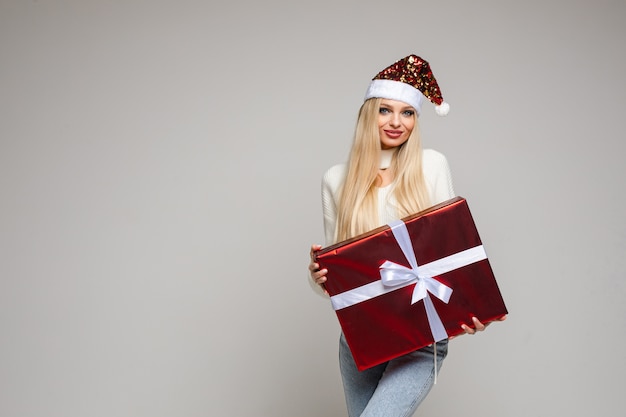 Handsome caucasian woman with blonde hair and pretty face in warm sweater, jaens and christmas hat holds a big red box