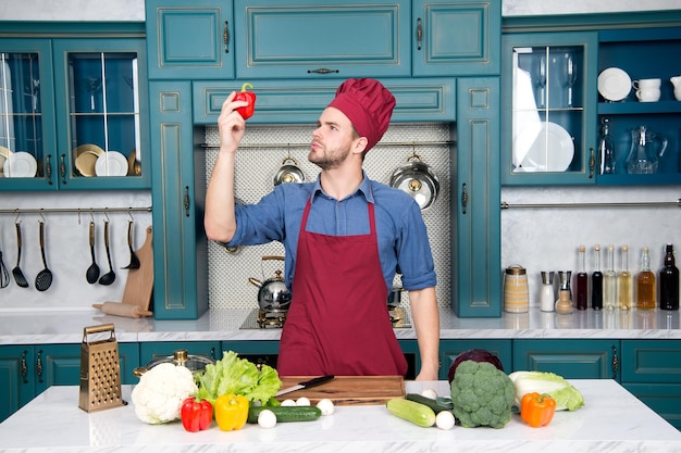 豪華な台所用品でいっぱいの木製の表面を持つキッチンで準備している家庭で野菜を調理しているテーブルでエプロンのハンサムな白人の若い男