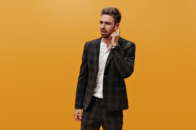 Handsome charming bearded young man in checkered suit and white shirt poses on orange wall and touches wireless headphones.