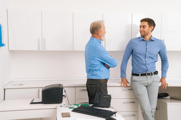 Handsome cheerful adult manager and smiling young colleague having fun conversation in office Handsome young man talking with presumed elderly father before genetic paternity test at medical clinic