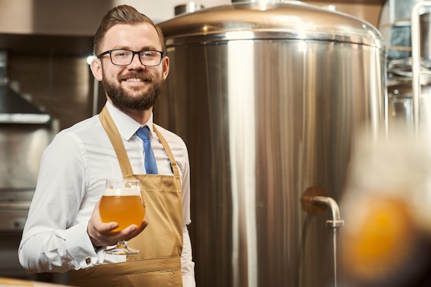 Handsome, cheerful brewer in white shirt and brown apron standing, holding cold beer glass with foam. Positive professional smiling, posing on background of storage tank.