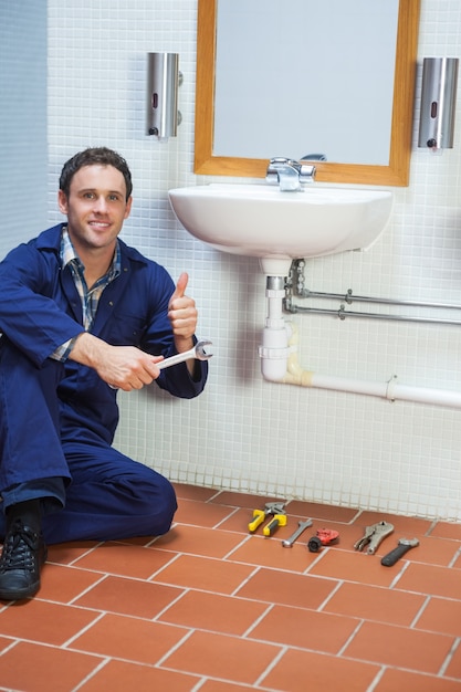 Handsome cheerful plumber sitting next to sink showing thumb up