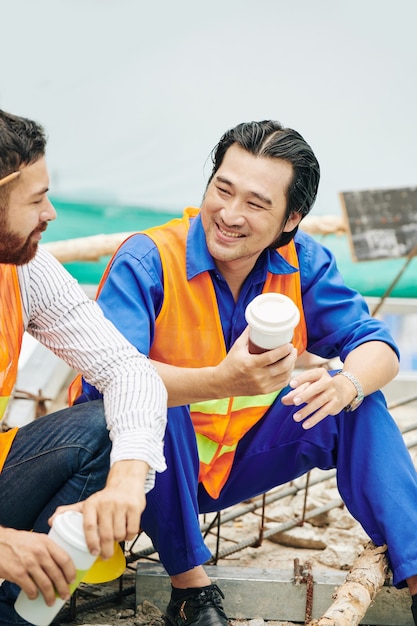 Handsome cheerful Vietnamese builder drinking coffee and talking to colleague during break