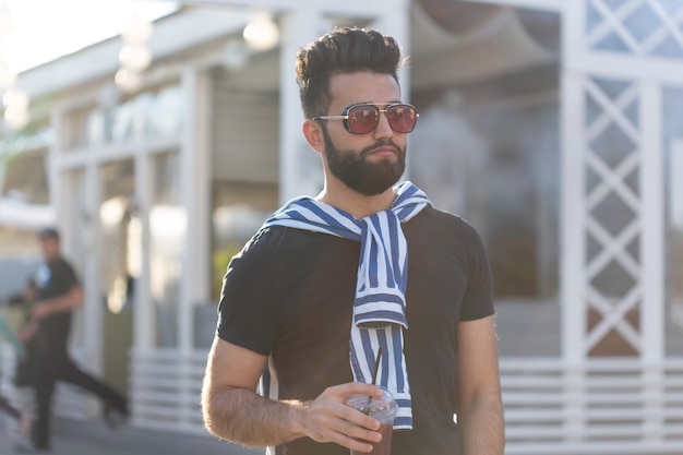 Handsome cheerful young arabian student with a stylish mustache and a beard with juice in his hands, walking around the city after a working day. Concept of positive and rest.