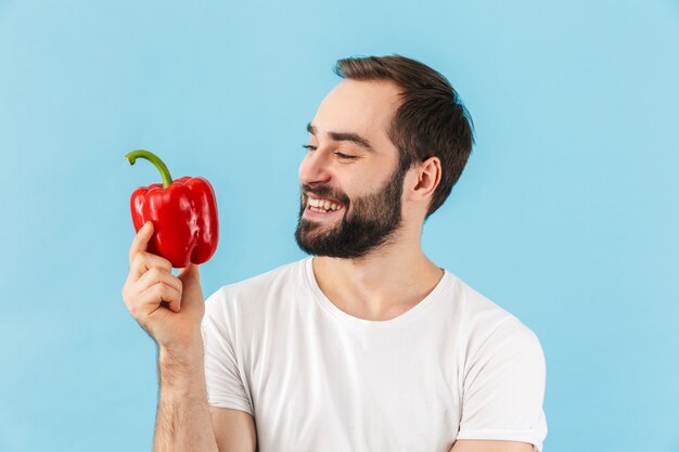 Handsome cheerful young bearded man wearing t-shirt standing isolated over blue wall, showing red capsicum