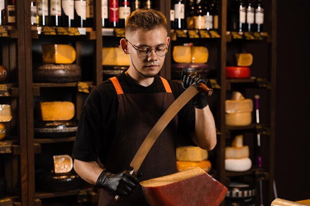 Handsome cheese sommelier with big knife for cut cheese bearded\
hipster in fashion glasses working in cheese shop creative idea for\
advertising