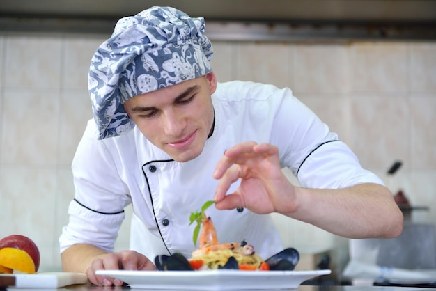 Handsome chef dressed in white uniform decorating pasta salad\
and seafood fish in modern kitchen