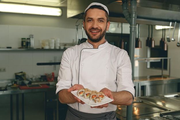 Foto bello chef del ristorante japanies che mostra piatto con sushi in piedi sulla cucina