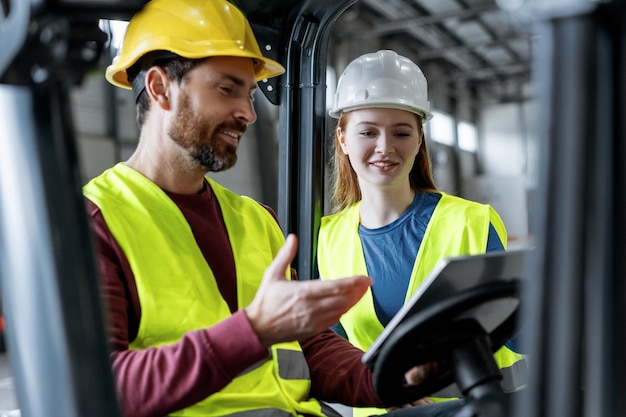 Handsome colleagues in helmets and communicate man driving forklift using digital tablet