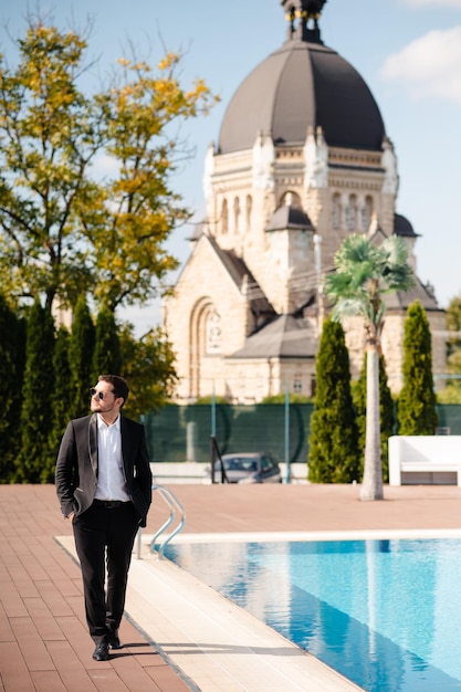 Photo handsome concevied young groom man in suit and sunglasses walking near swimming pool at sunny day church and nature on the background wedding day