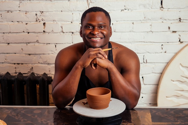 Handsome and confident afro american man potter bare body apron naked sculpts clay vase sexy sculptor african student In workshop on the background of a rack with pottery dishes art do it yourself