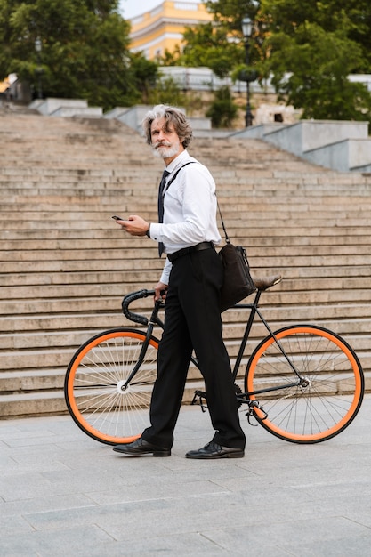 Bello fiducioso uomo d'affari barbuto che indossa tuta, porta una valigetta camminando all'aperto con la bicicletta, usando il telefono cellulare