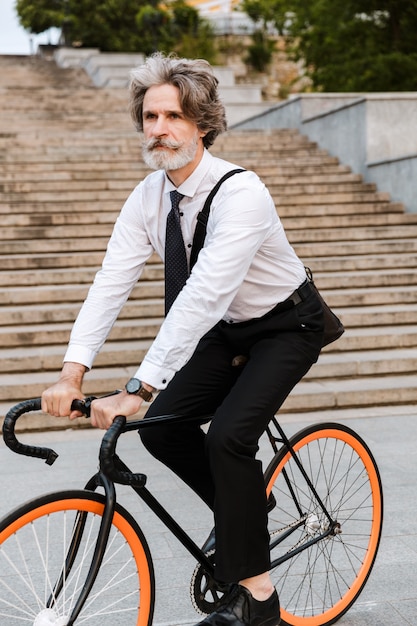 Handsome confident bearded man wearing suit, carrying briefcase outdoors riding on a bicycle