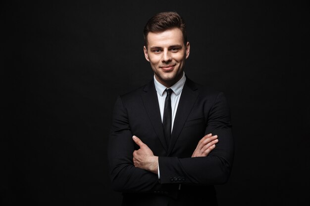 Handsome confident businessman wearing suit standing isolated over black wall, arms folded