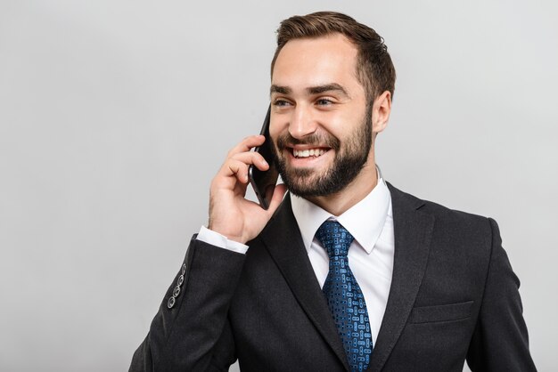 Un bell'uomo d'affari fiducioso che indossa un abito in piedi isolato su un muro grigio, parlando al telefono cellulare