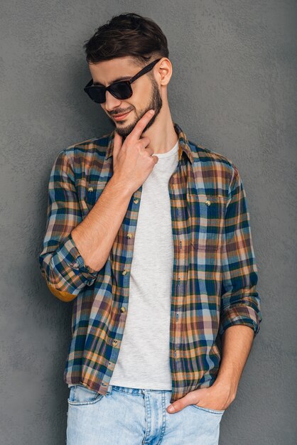 Handsome and confident. Cheerful young man in sunglasses holding hand on chin and looking down with smile while standing against grey background