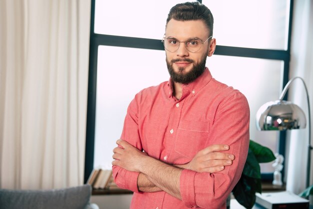 Handsome confident and happy bearded man in eyeglasses and shirt is standing with crossed arms and looking on the camera