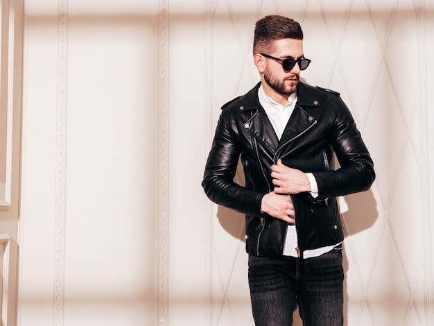 Handsome confident model sexy stylish man in biker leather\
jacket and black jeans fashion hipster male posing near white wall\
in studio interior at sunny day shadow from window in\
sunglasses