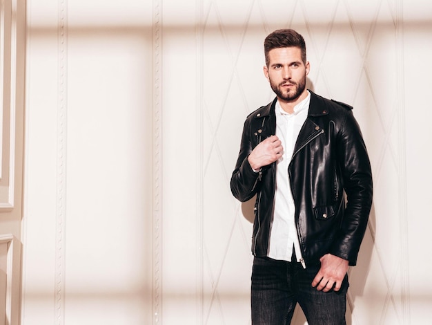 Handsome confident model sexy stylish man in biker leather
jacket and black jeans fashion hipster male posing near white wall
in studio interior at sunny day shadow from window