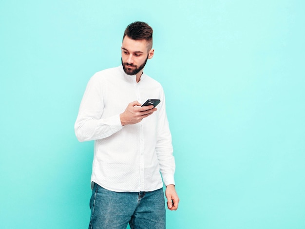 Modello bello e sicuro di sé uomo sexy ed elegante vestito con camicia e jeans moda uomo hipster in posa vicino al muro blu in studio holding smartphone guardando lo schermo del cellulare usando le app