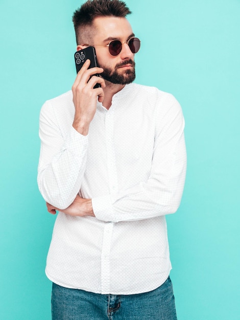 Handsome confident modelSexy stylish man talking at smartphone Fashion hipster male posing near blue wall in studio Holding phone With cellphone In sunglasses Isolated