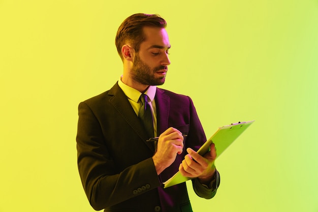 Handsome confident young businessman wearing formal suit standing isolated over yellow wall, holding notepad