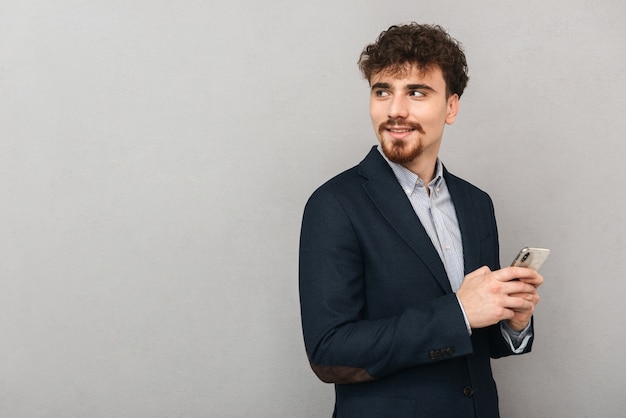 Handsome confident young businessman wearing a jacket standing isolated over gray, holding mobile phone