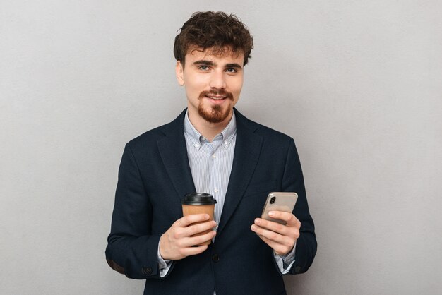 Красивый уверенный молодой бизнесмен в куртке, стоящей изолированно над серым, используя мобильный телефон, держа чашку кофе на вынос