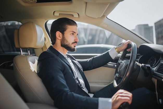 ハンサムで自信を持って若い男は車に座って、まっすぐに見えます。彼は車を運転します。内部は豪華に見えます。彼は片手でハンドルを握り、もう片方をハンドブレーキで握ります。