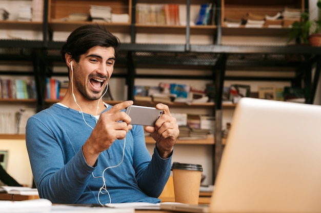 Handsome confident young man wearing earphones sitting at the library desk, working/studying, holding mobile phone