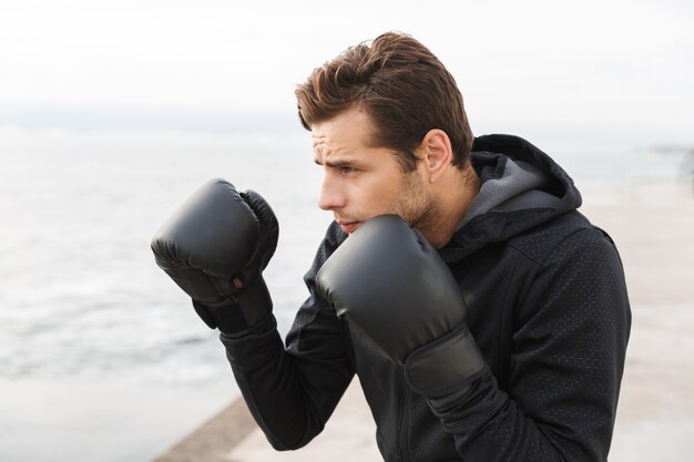 Handsome confident young sportsman working out at the beach, doing boxing exercises