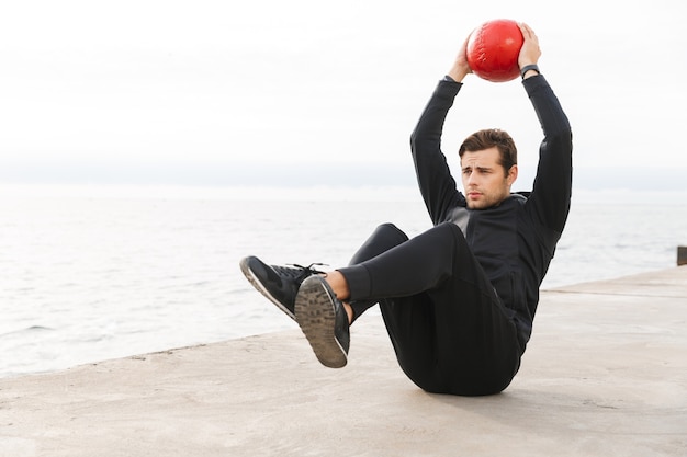 Handsome confident young sportsman working out at the beach, doing exercises with a heavy ball