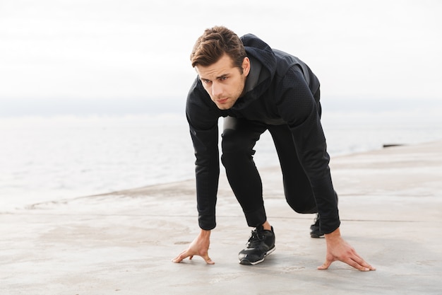 Handsome confident young sportsman working out at the beach, ready to start running, posing