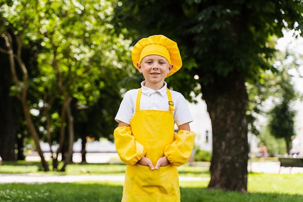 黄色いシェフの帽子とエプロンの黄色い制服を着たハンサムな料理人の子供が屋外でポーズをとる食料品店やカフェのクリエイティブな広告