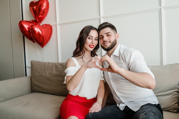 Handsome couple man and woman making heart shape with hands with red balloons sitting at home on couch